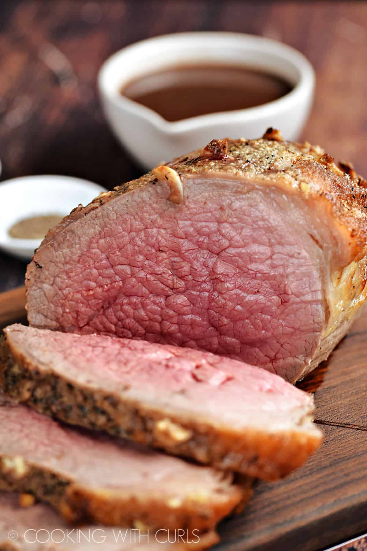 Medium-rare roast beef sliced on a dark wood cutting board with a bowl of gravy in the background.