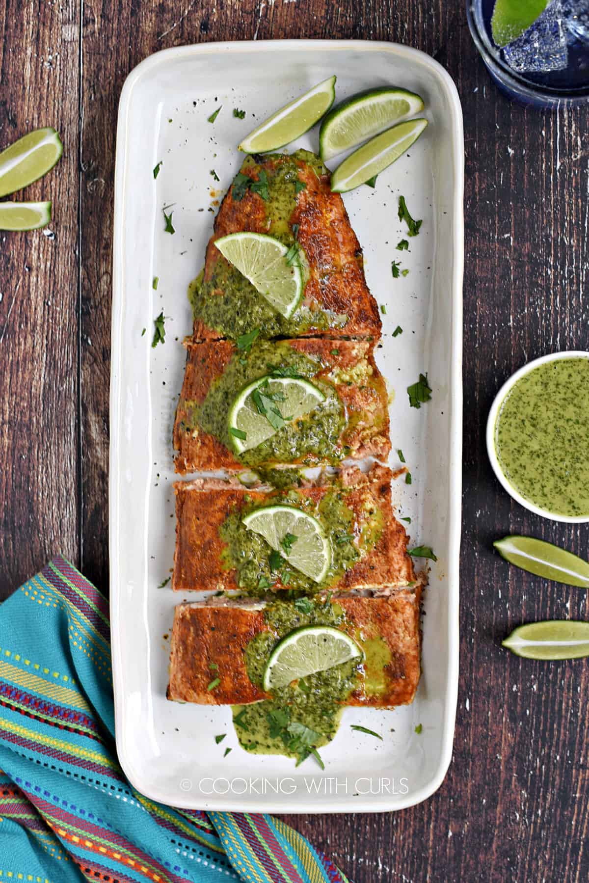 Looking down on a baked salmon filet topped with cilantro sauce and lime slices on a white platter with a bowl of sauce on the side.