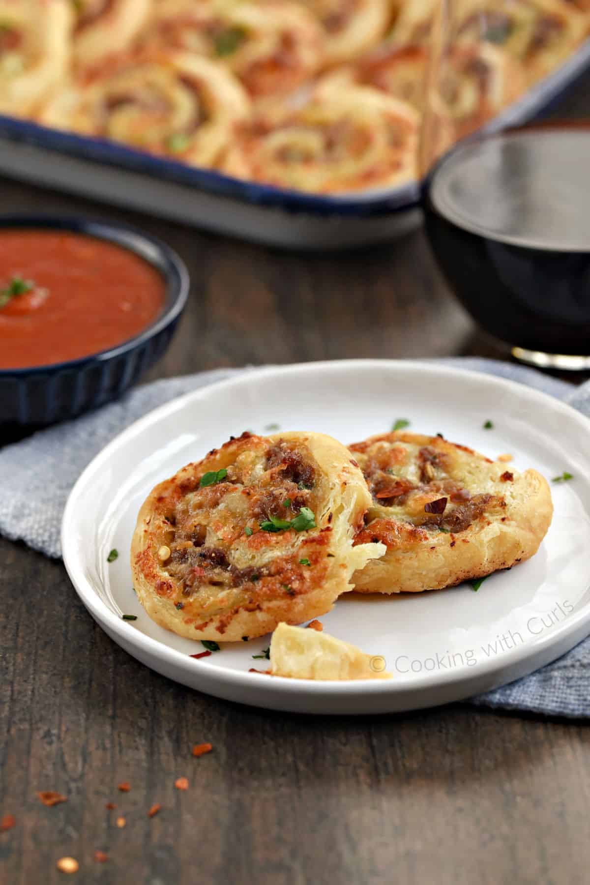 Two sausage pinwheels with puff pastry sitting on a small plate with pizza sauce in the background.