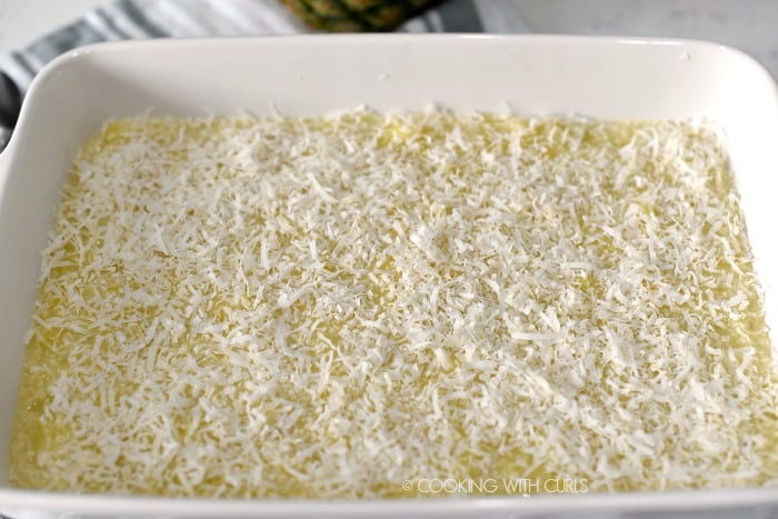 Shredded coconut sprinkled over the pineapple filling in a white baking dish. 