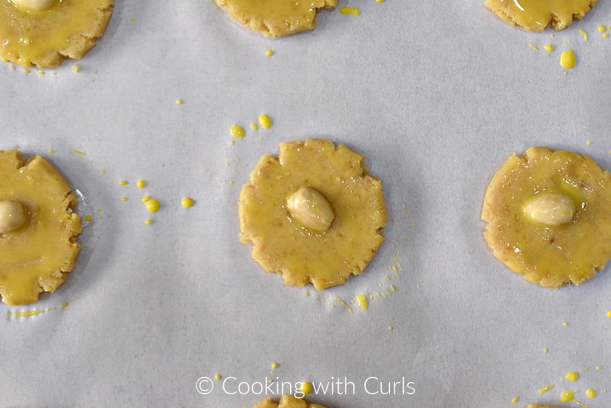 Six flattened cookies topped with a blanched almond and egg wash on parchment paper. 