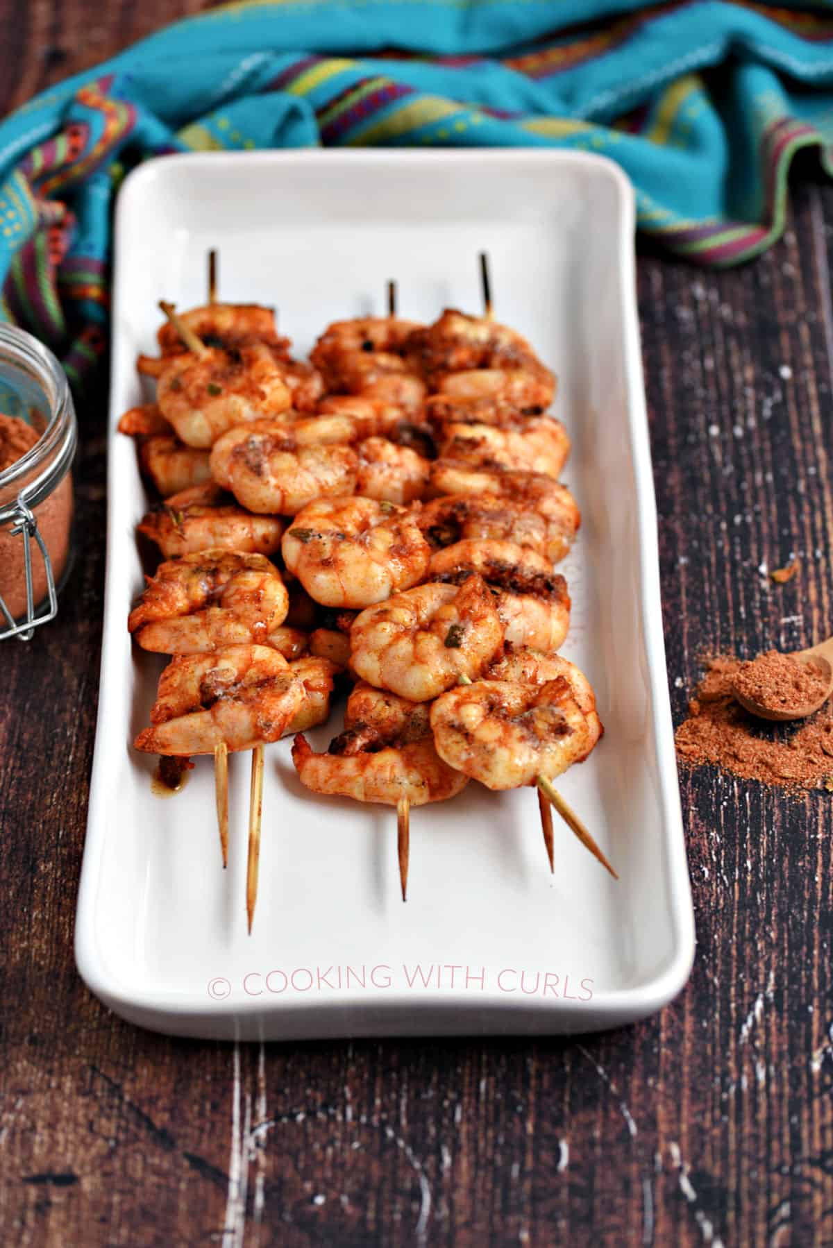Skewered shrimp stacked on a white platter with Mexican seasoning in the background. 