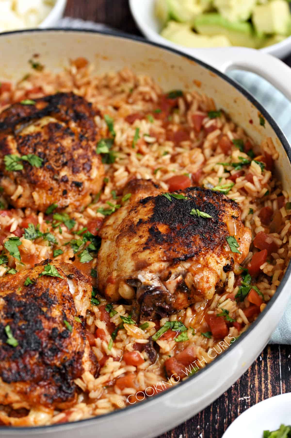 Seasoned chicken thighs and Spanish rice in a white skillet with chopped avocado and queso cheese in the background.