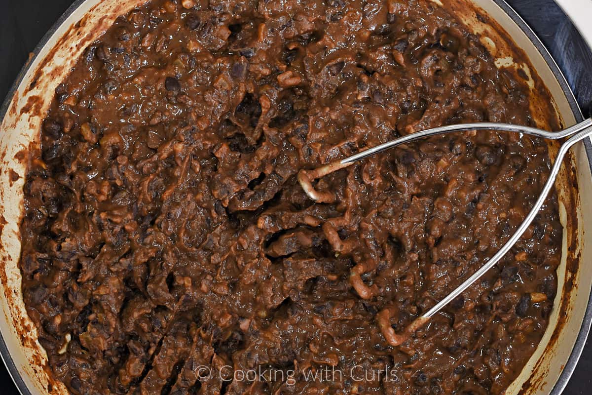 Smashed Refried Black Beans in a skillet with a potato masher on the side. 