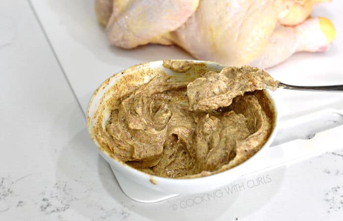 Softened butter mixed with southwest seasoning in a small white bowl with a whole chicken on a white cutting mat in the background. 