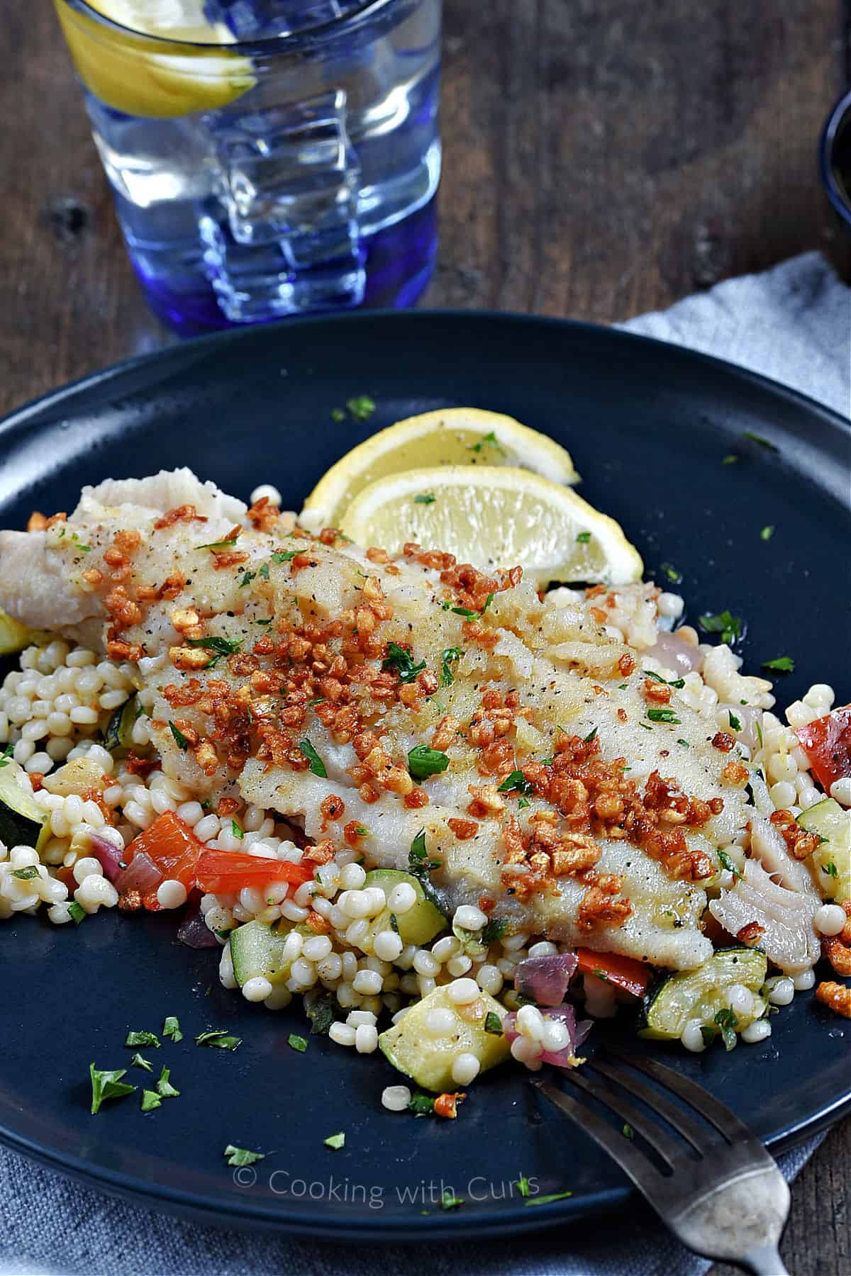 Sole with Caramelized Garlic on a bed of couscous with vegetables and lemon wedges on a dinner plate.