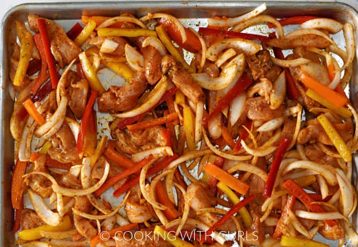 Strips of marinade covered chicken, onion, red, orange and yellow bell peppers on a sheet pan. 