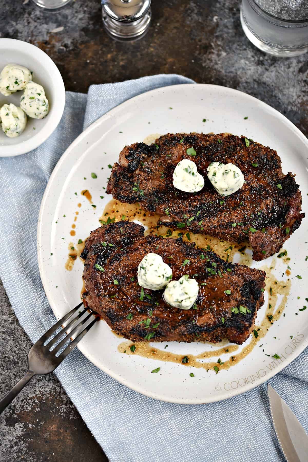 Looking down on two herb butter topped ribeye steaks on a white dinner plate with a bowl of butter hearts in the upper left corner.