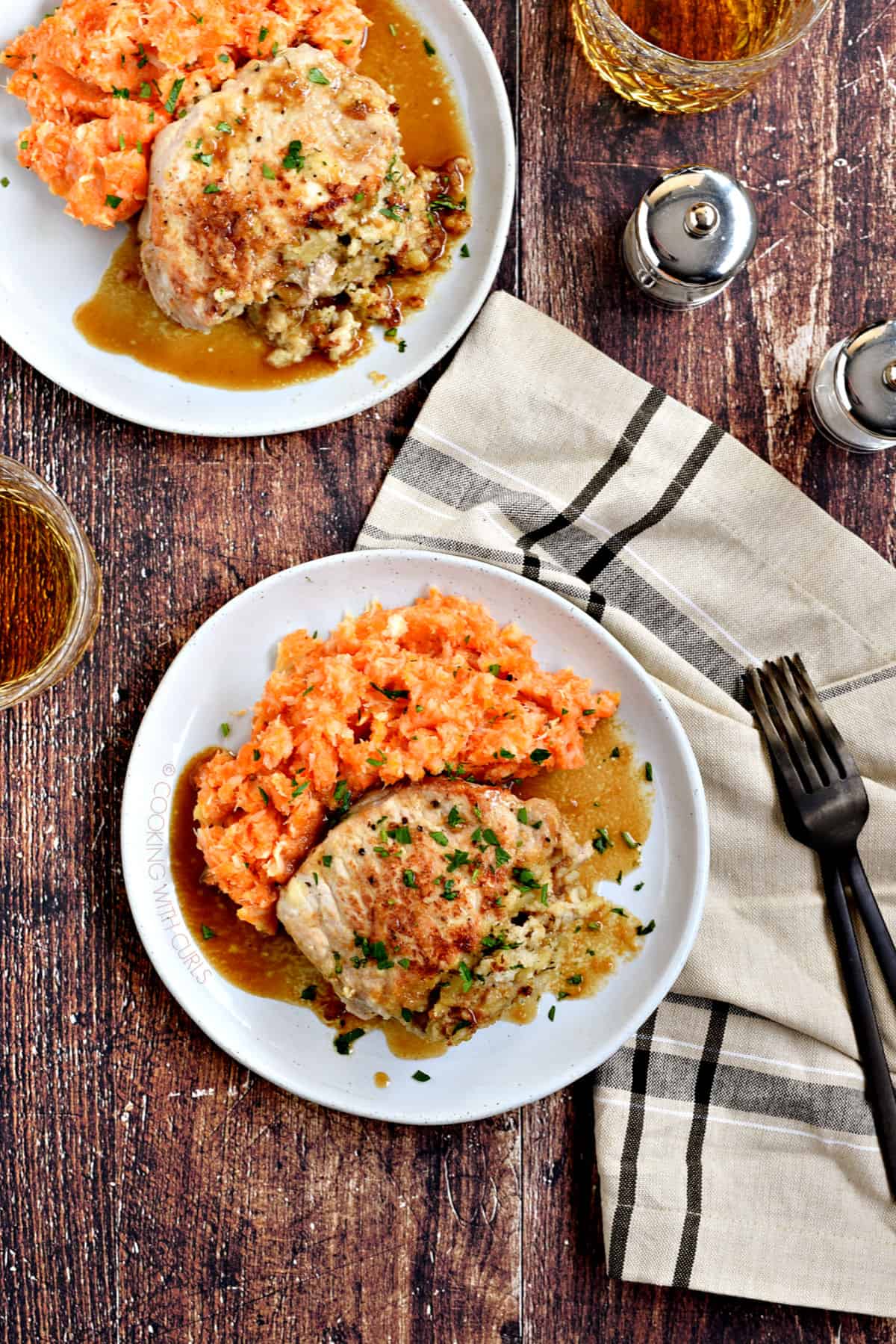 Looking down on two plates of A stuffed pork chop covered in an apple-bourbon sauce and served with carrot parsnip mash with a glass of whisky in the background.