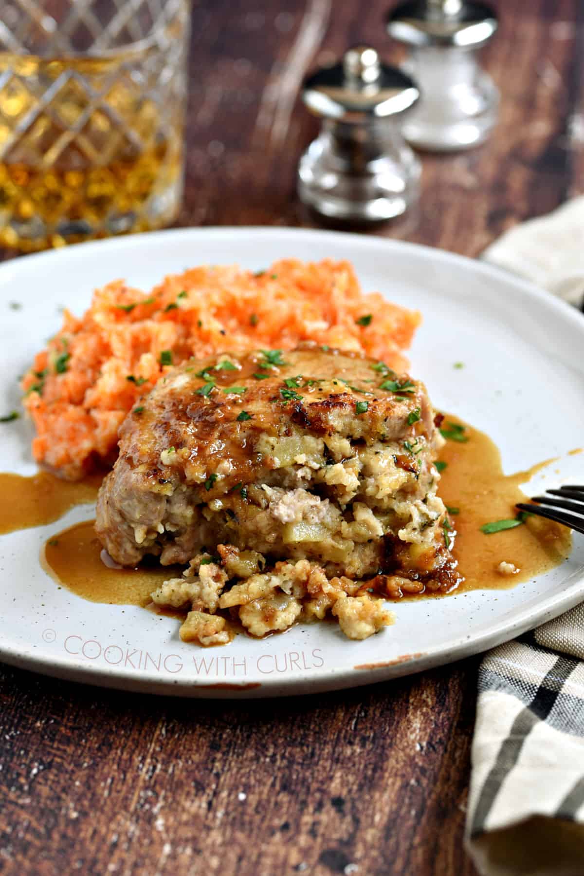 A stuffed pork chop covered in an apple-bourbon sauce and served with carrot parsnip mash on a large white plate with a glass of whisky in the background.