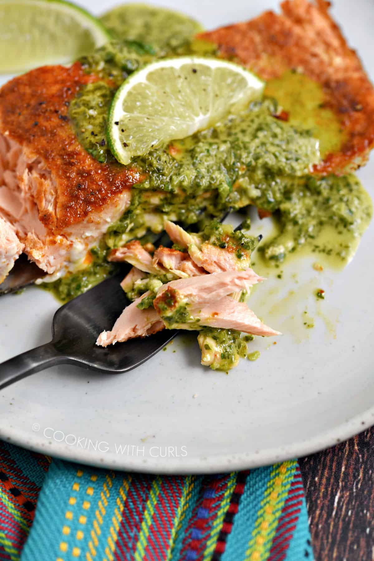 Flaky salmon on a black fork with a salmon filet topped with cilantro sauce and lime slice on a white plate.