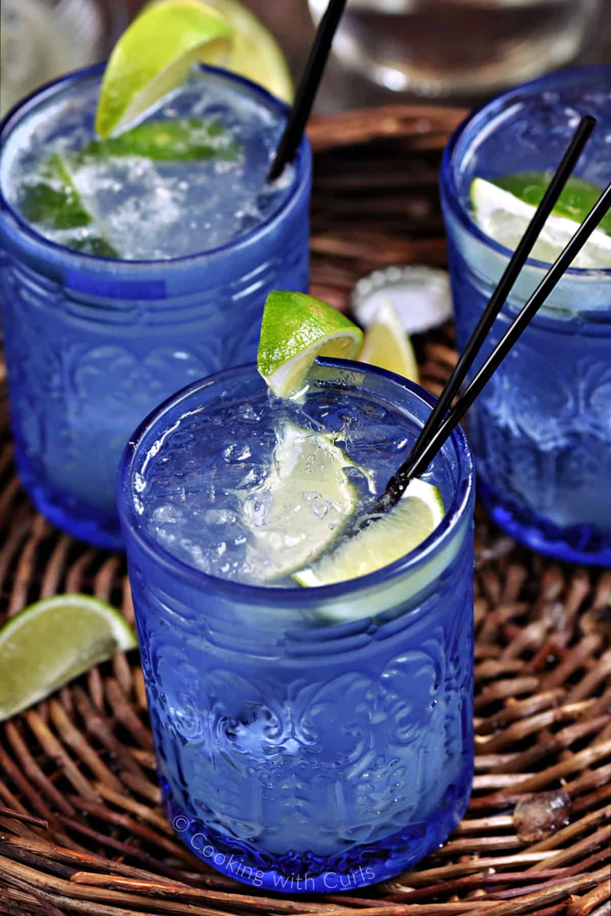 Three blue glasses filled with ice, tequila, lime juice and mineral water with lime wedge garnish. 