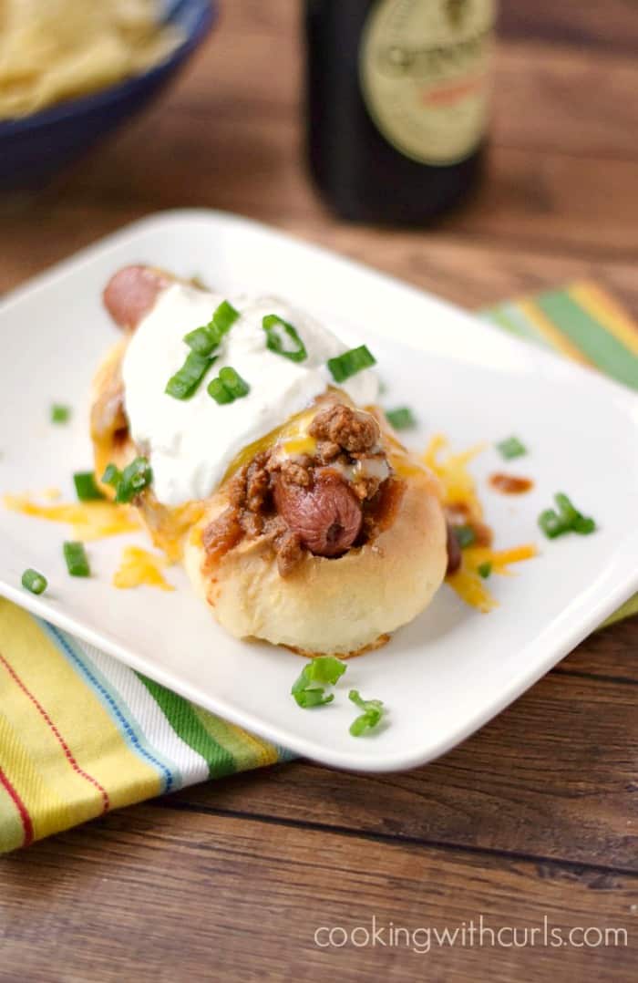 a hot dog smothered in chili and melted cheese, topped with sour cream and sliced green onions sitting on a white plate with a bottle of Guinness in the background