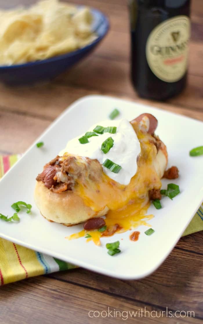 a chili and melted cheese covered hot dog in a homemade hot dog bun sitting on a white plate with a bottle of Guinness in the background