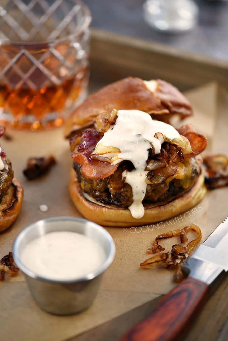 A burger topped with grilled onions, bacon, and whiskey mayonnaise on a pretzel bun with a glass of whiskey in the background.