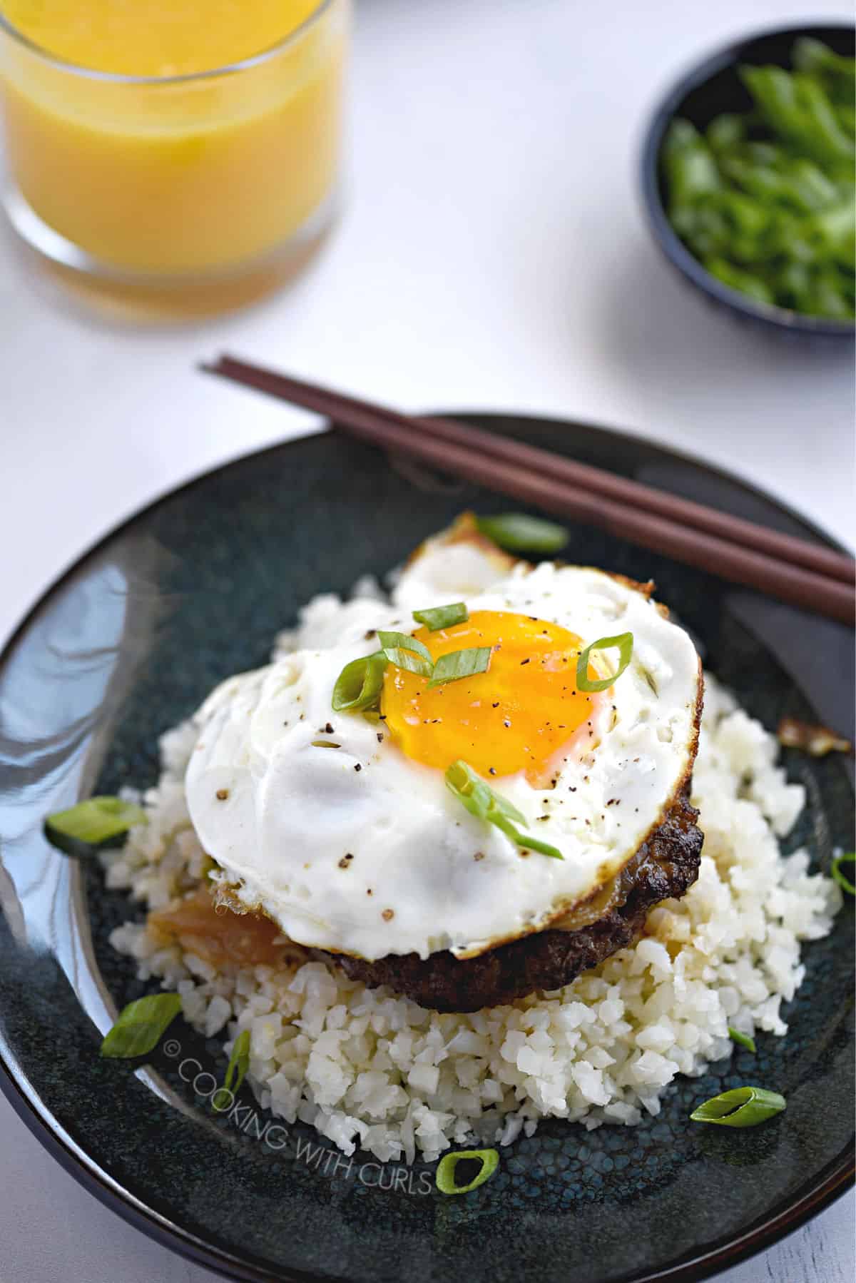 looking down on a sunny-side up egg on top of a burger patty covered in gravy on a bed of rice with a glass of orange juice and small bowl of green onions in the background.