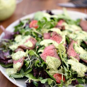 A large white plate topped with salad greens, sliced steak, cherry tomatoes and cilantro-lime dressing