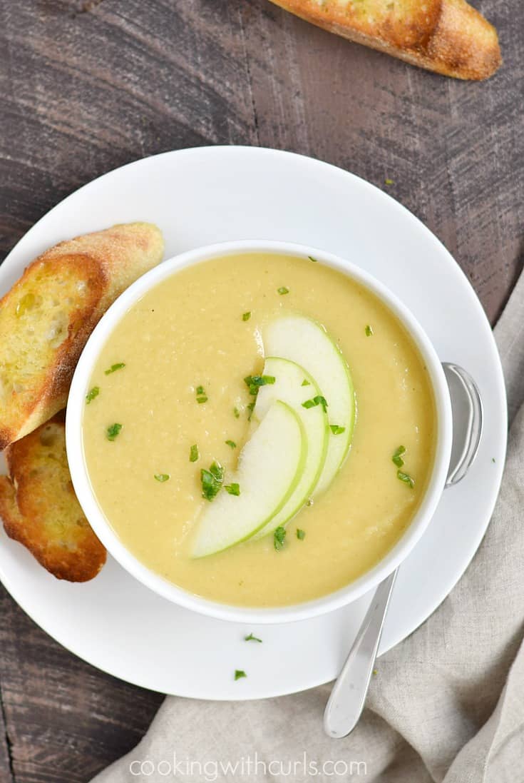 Looking down on creamy parsnip and apple soup in a white bowl topped with green apple slices and green onions with toasted bread slices on the side.