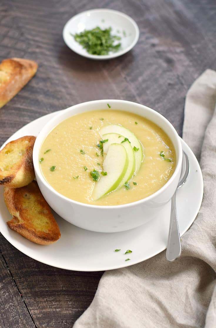 Creamy parsnip and apple soup in a white bowl sitting on a white plate topped with green apple slices and chopped green onions with toasted bread slices and a bowl of green onions in the background.