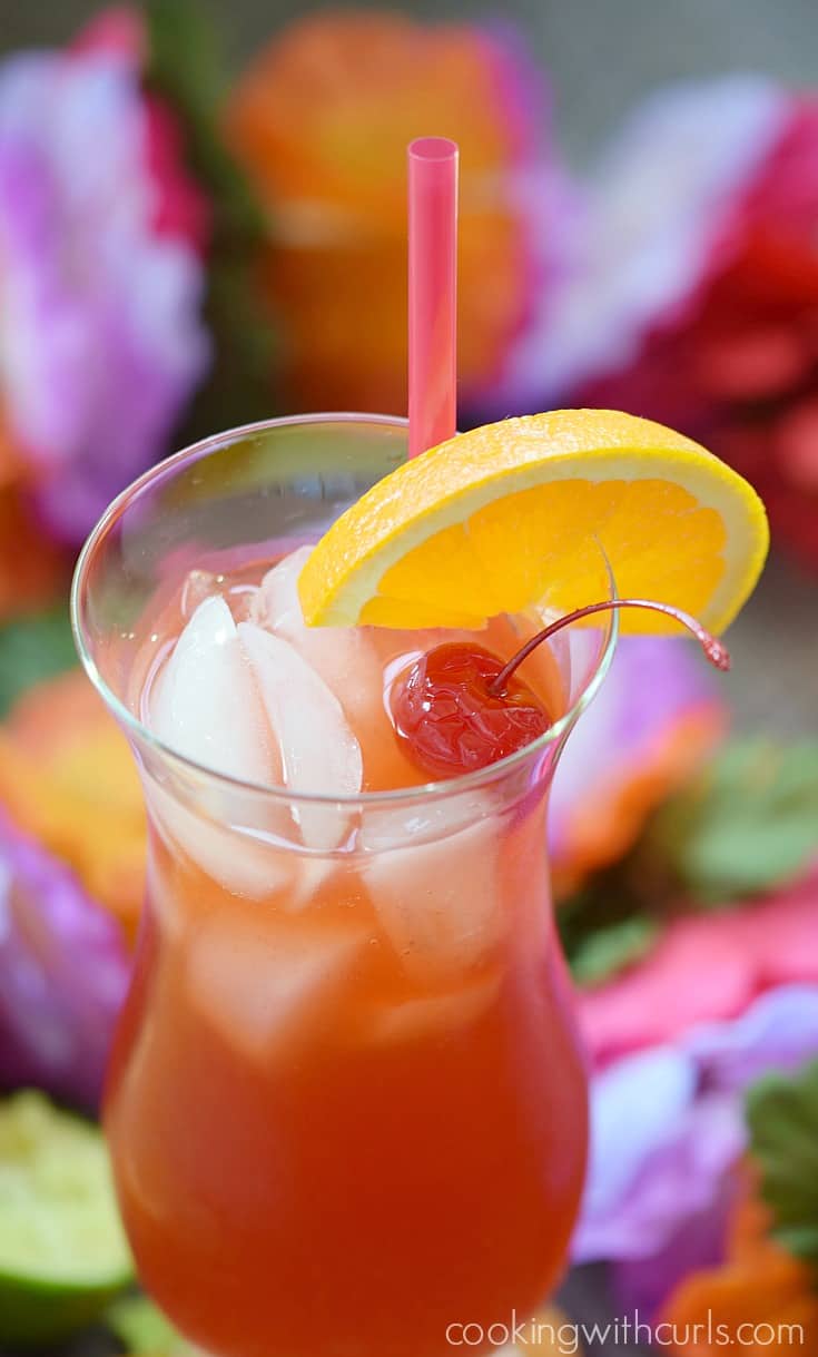 Bright red cocktail in a hurricane glass garnished with orange wheel and cherry.