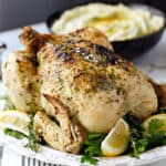 An Instant Pot Whole Greek Chicken on a white platter with thyme and lemon wedges, with a bowl of mashed potatoes in the background and title across the top.
