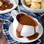 Chocolate Gravy overflowing a white gravy boat sitting on a white plate with a ladle sticking up out of the backside, with a place of chocolate gravy covered biscuits on the left and a bowl of biscuits in the upper right corner and title graphic across the bottom.