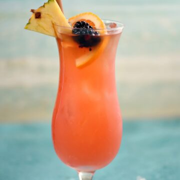 a pink cocktail in an ice filled hurricane glass garnished with a pineapple wedge, orange slice, blackberry and bamboo straw with an ocean image in the background.