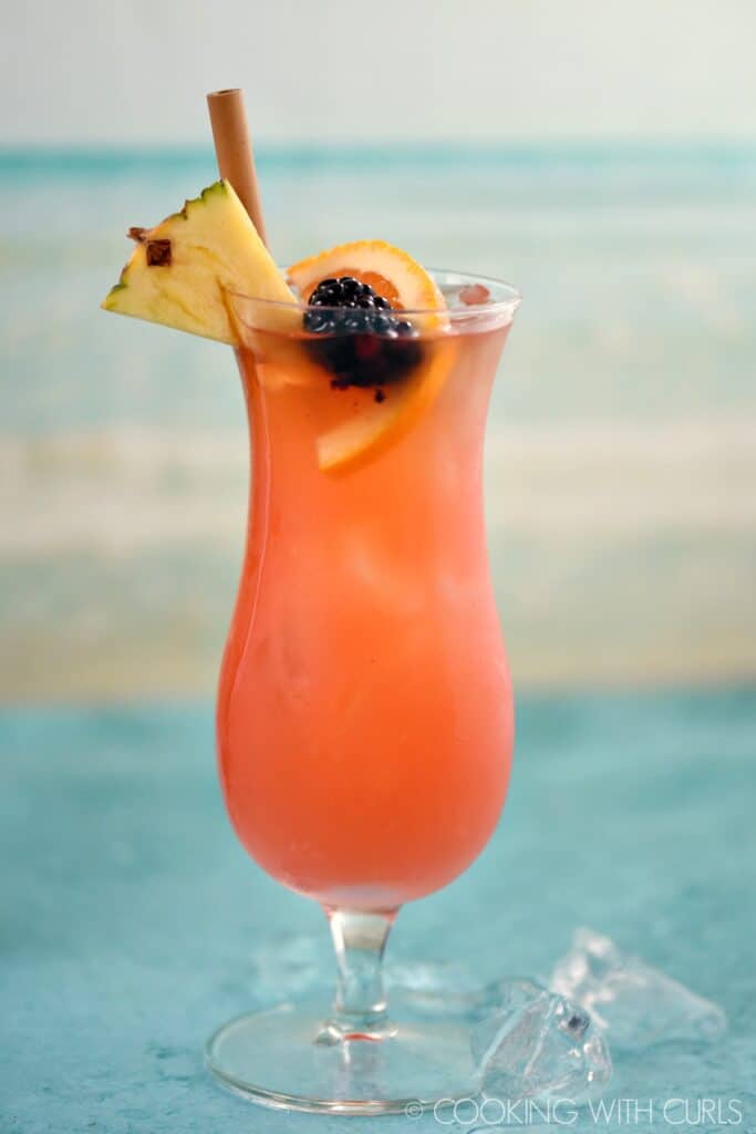 an ice filled hurricane glass with a pink cocktail that is garnished with a pineapple wedge, orange slice, blackberry and bamboo straw.