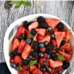 Looking down on a white serving bowl filled with watermelon wedges, blackberries, strawberries and blueberries with title across the top of the image.