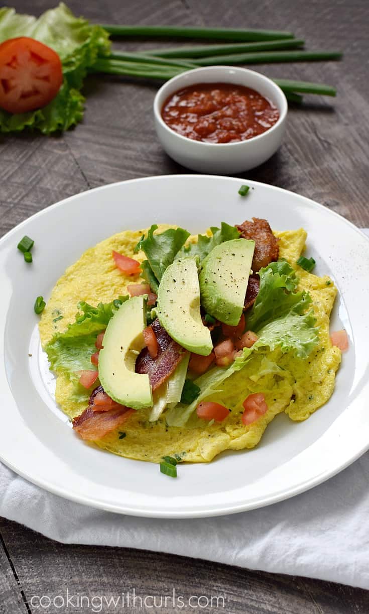 Avocado slices, bacon, chopped tomatoes, and lettuce leaves on top of a scrambled egg "tortilla" on a small plate.