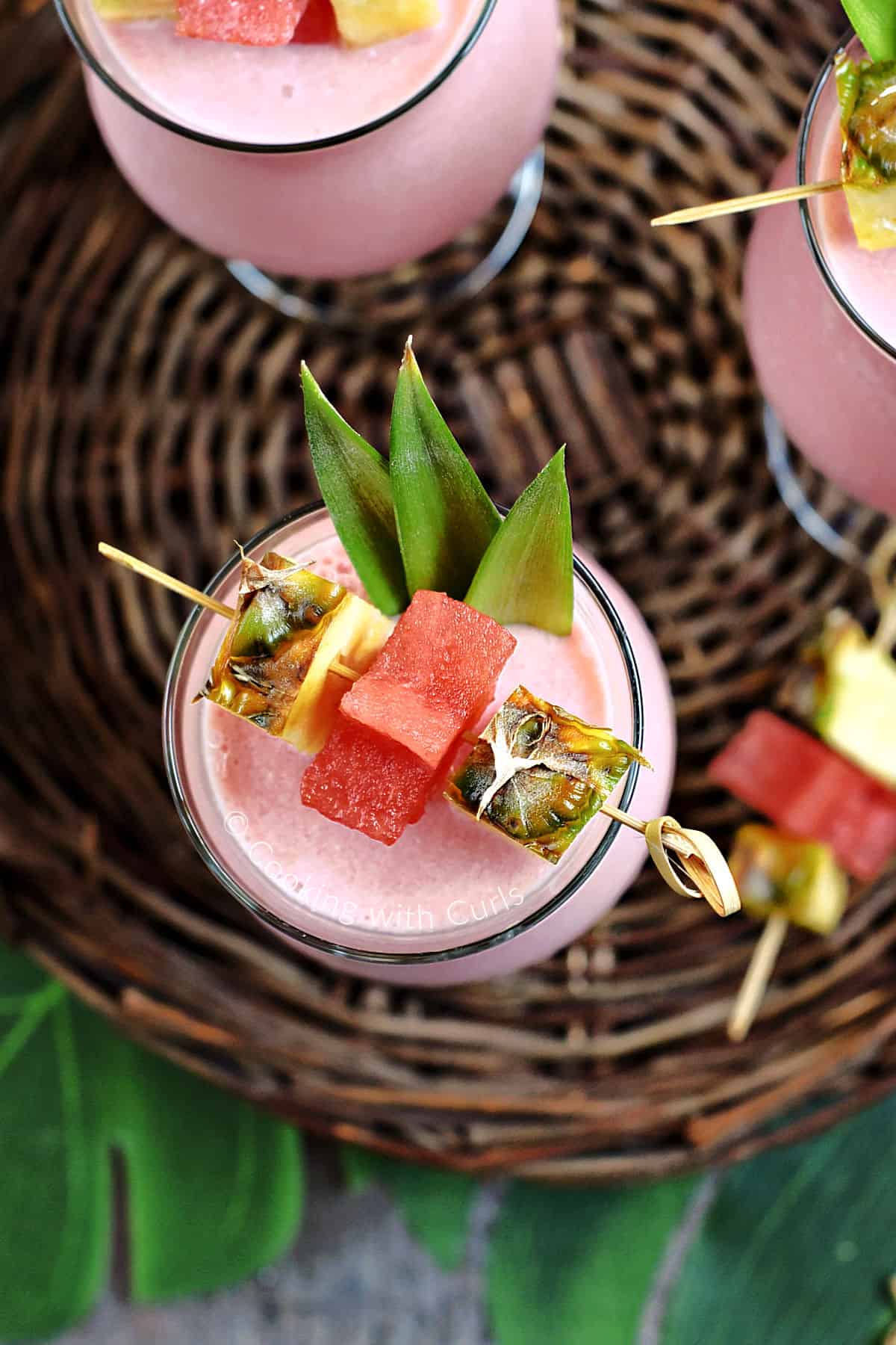 Three tropical frozen watermelon pina colada drinks on a rattan tray garnished with pineapple, watermelon stars, and pineapple leaves. 