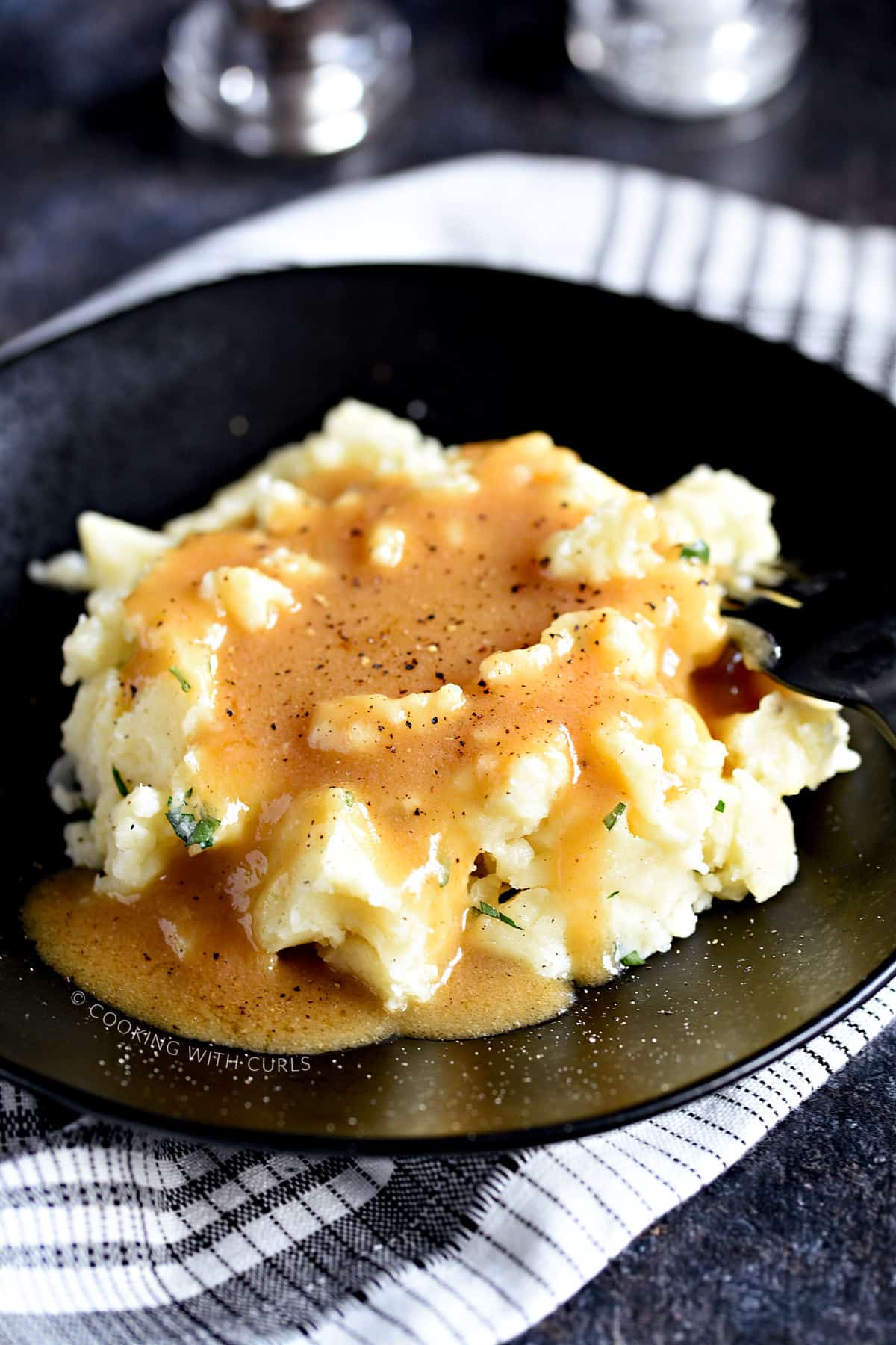 Turkey gravy poured over mashed potatoes and sprinkled with black pepper on a black plate that is sitting on a black and white napkin.