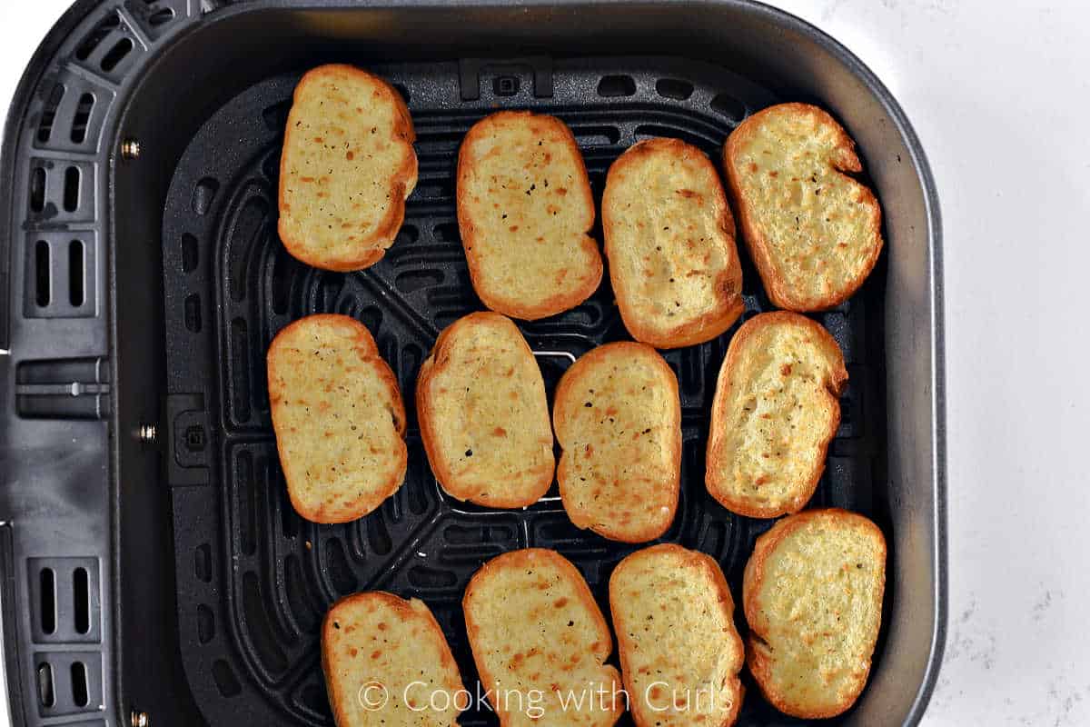 Twelve toasted baguette slices in the basket of an air fryer. 