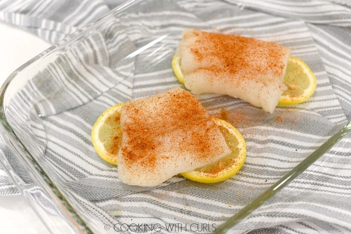two cod fillets laying on lemon slices in a glass baking dish sprinkled with southwest seasoning.