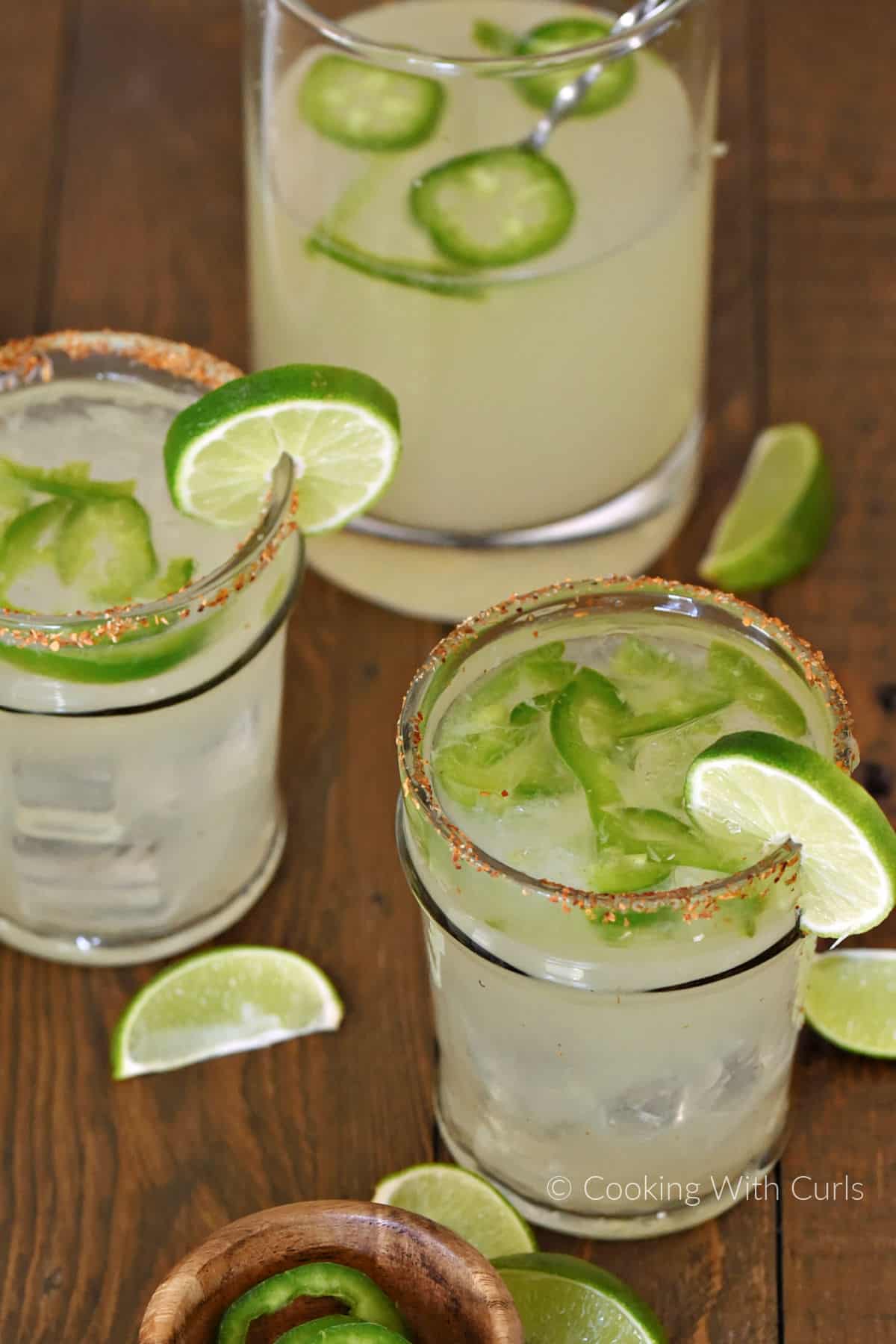 Two ice filled glasses and a pitcher with sliced jalapenos, margarita cocktail, and lime wheels. 