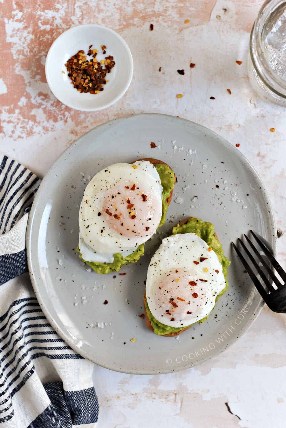 Two poached eggs sprinkled with flaked salt and red pepper flakes on top of two slices of avocado toast on a small plate. 