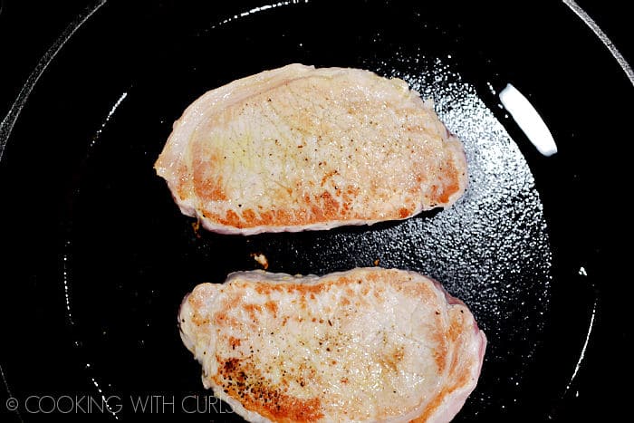 Two pork chops cooking in a cast iron skillet. 