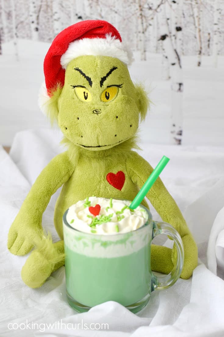 A Grinch doll sitting behind a clear glass mug filled with green hot chocolate topped with whipped cream, green sprinkles, and a red heart.