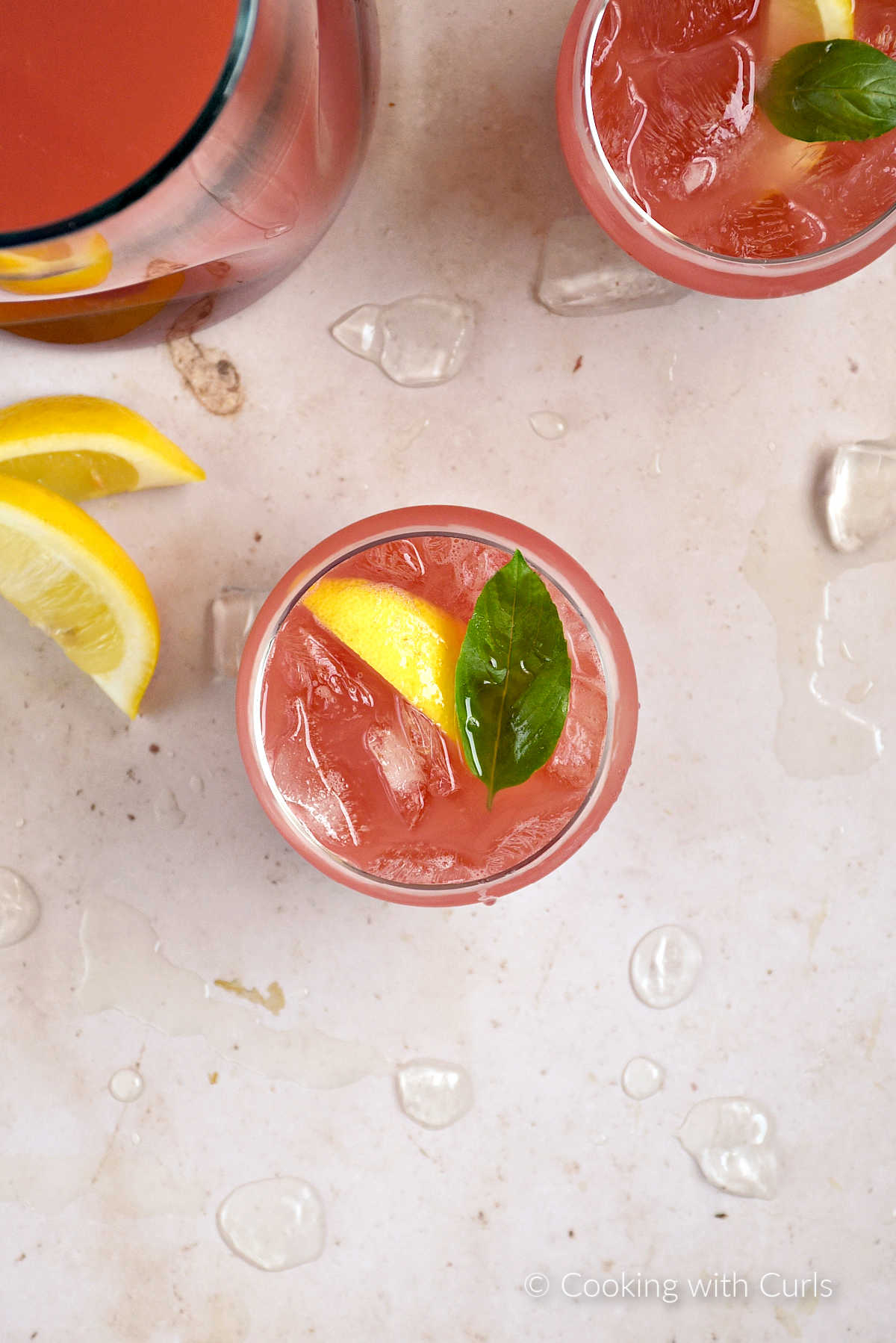 Looking down on a pitcher and two glasses of watermelon basil lemonade garnished with lemon wedge and basil leaf.