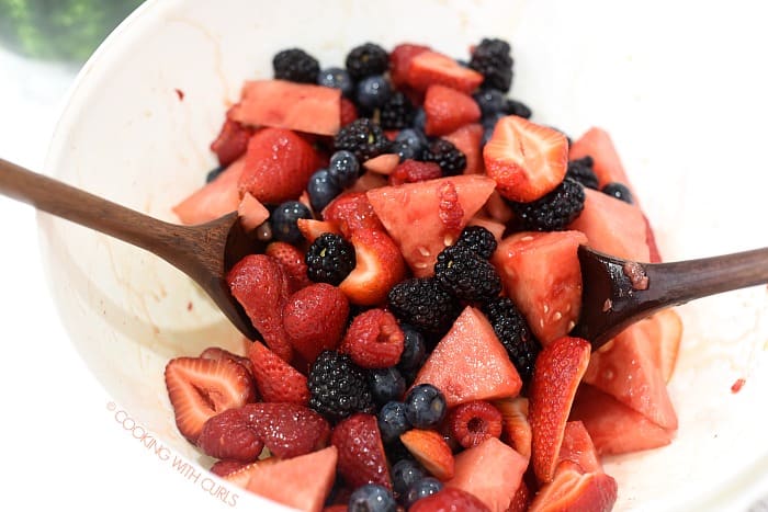 Watermelon, berries and honey citrus dressing tossed together with wooden spoons in a large white mixing bowl. 