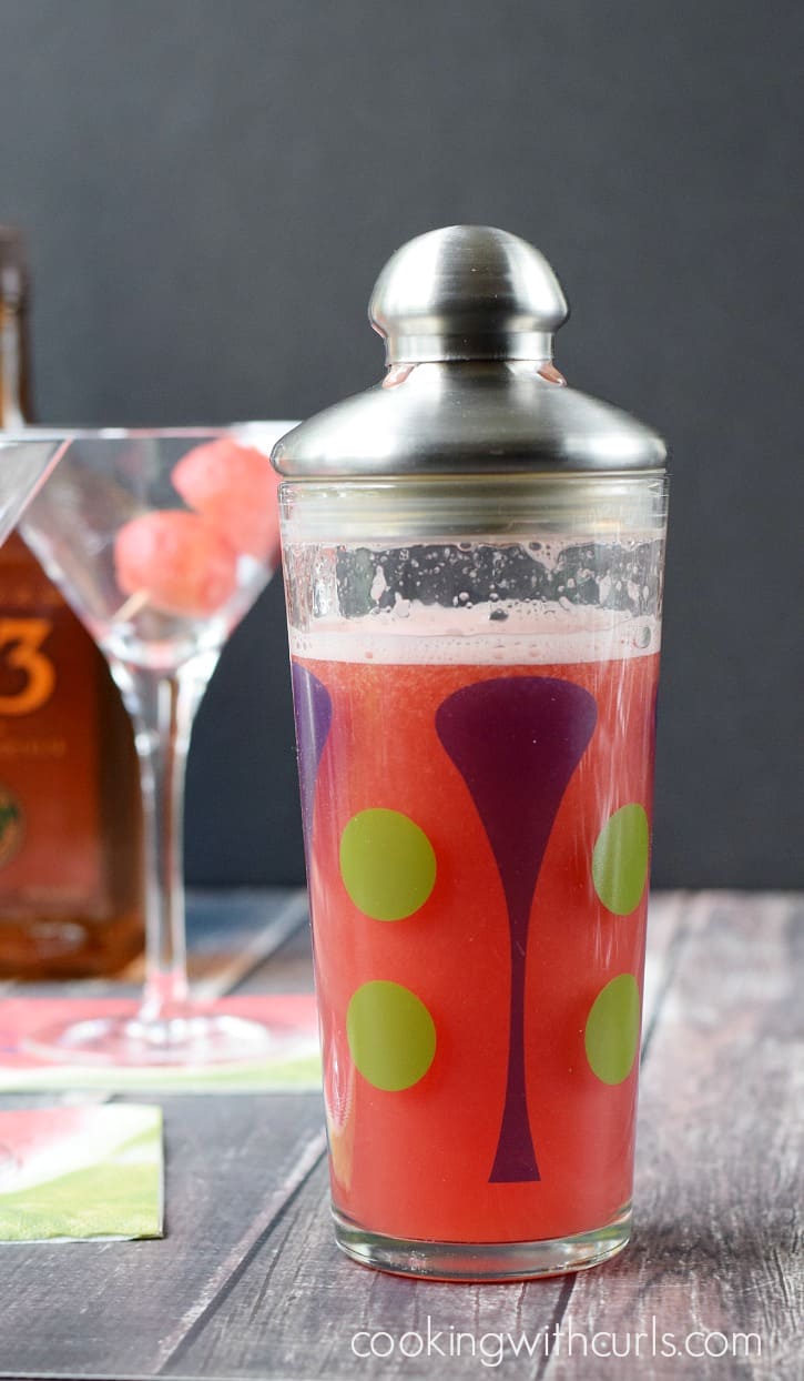 Watermelon drink in a cocktail shaker with an empty martin glass in the background with two watermelon balls on a toothpick.