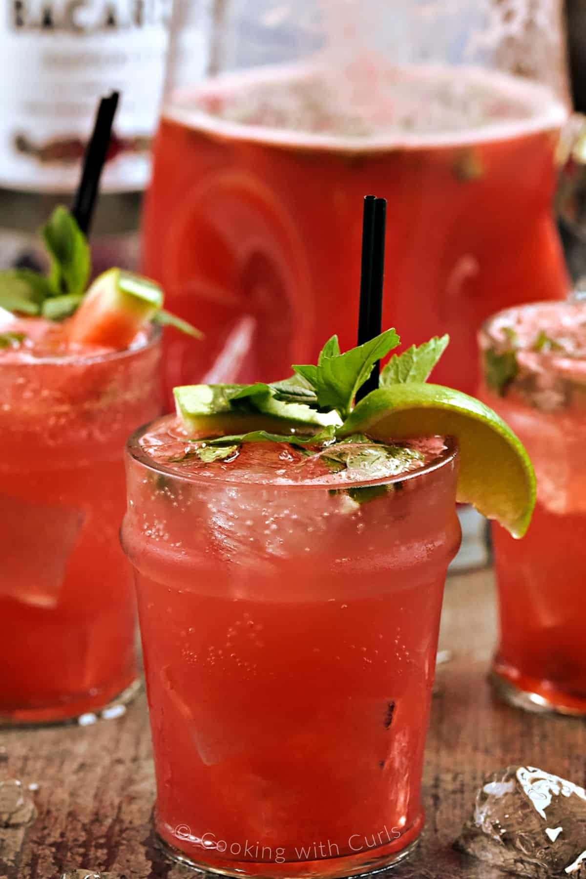 Three glasses and a pitcher filled with a bright pink cocktail garnished with watermelon and lime wedges with fresh mint sprigs.