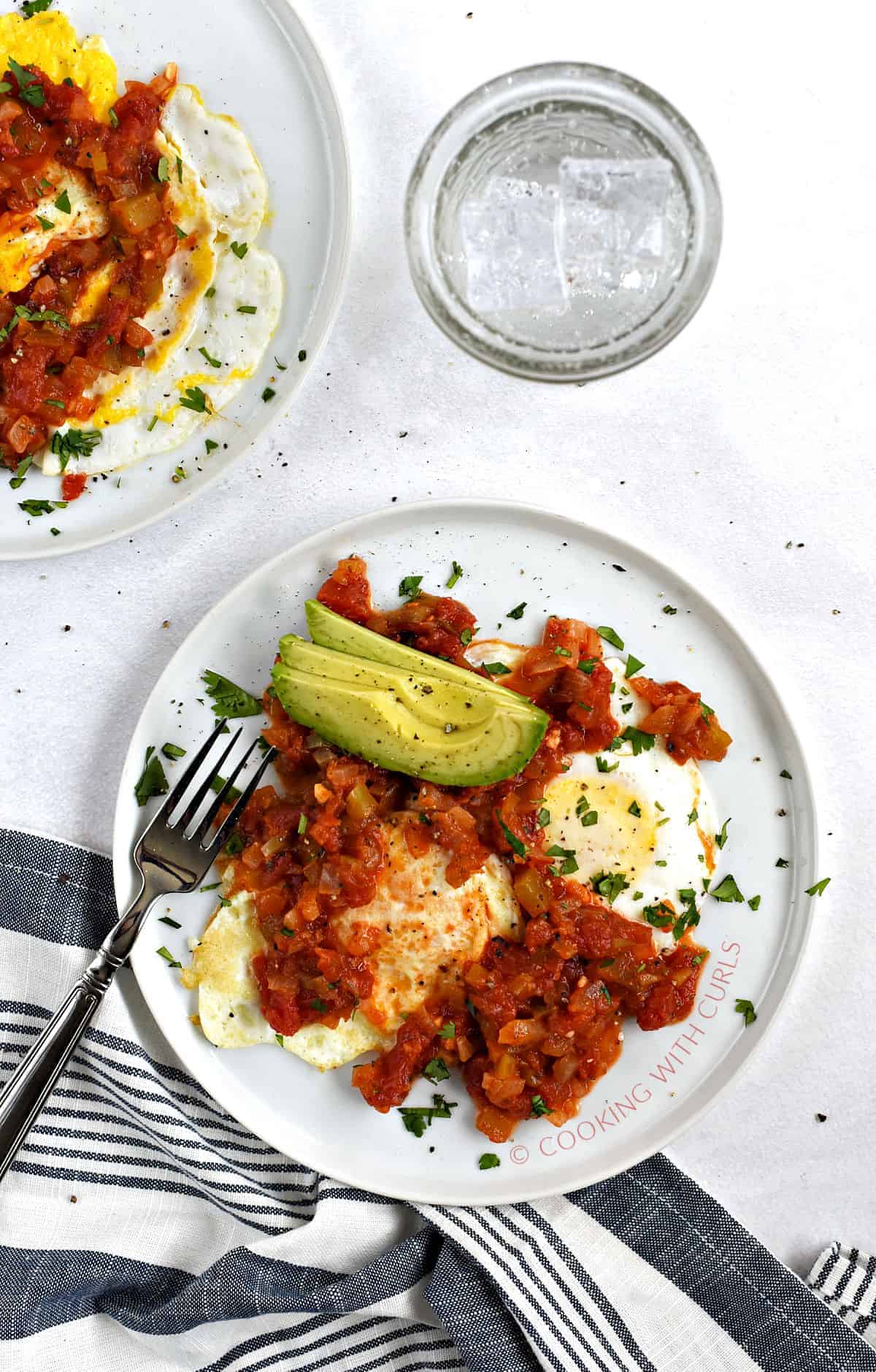 Two plates with two fried eggs, cooked tomato salsa, and sliced avocado.