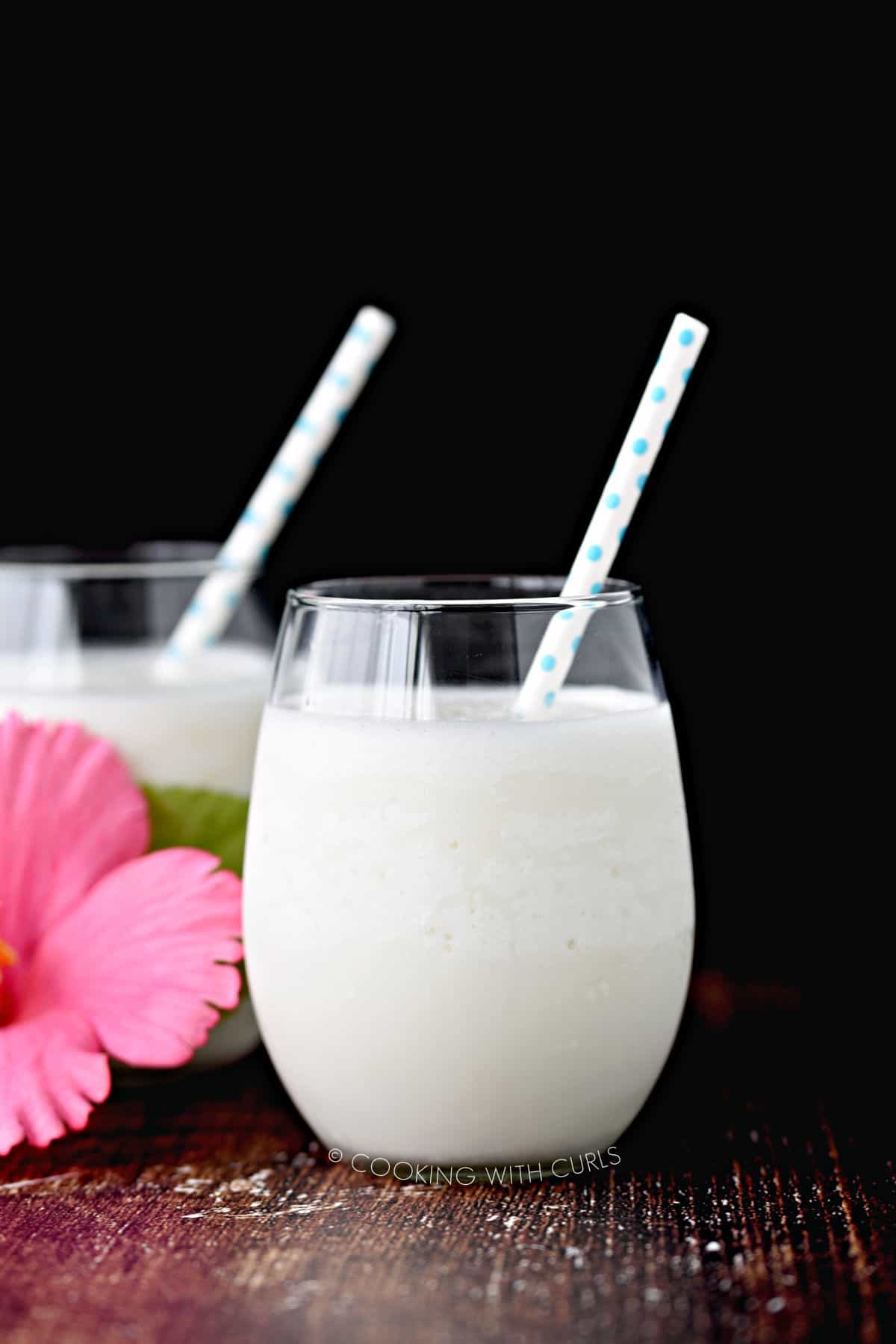 looking straight at two glasses filled with creamy cocktails, each with a blue and white polka dot straw and a pink flower sitting between the two glasses.