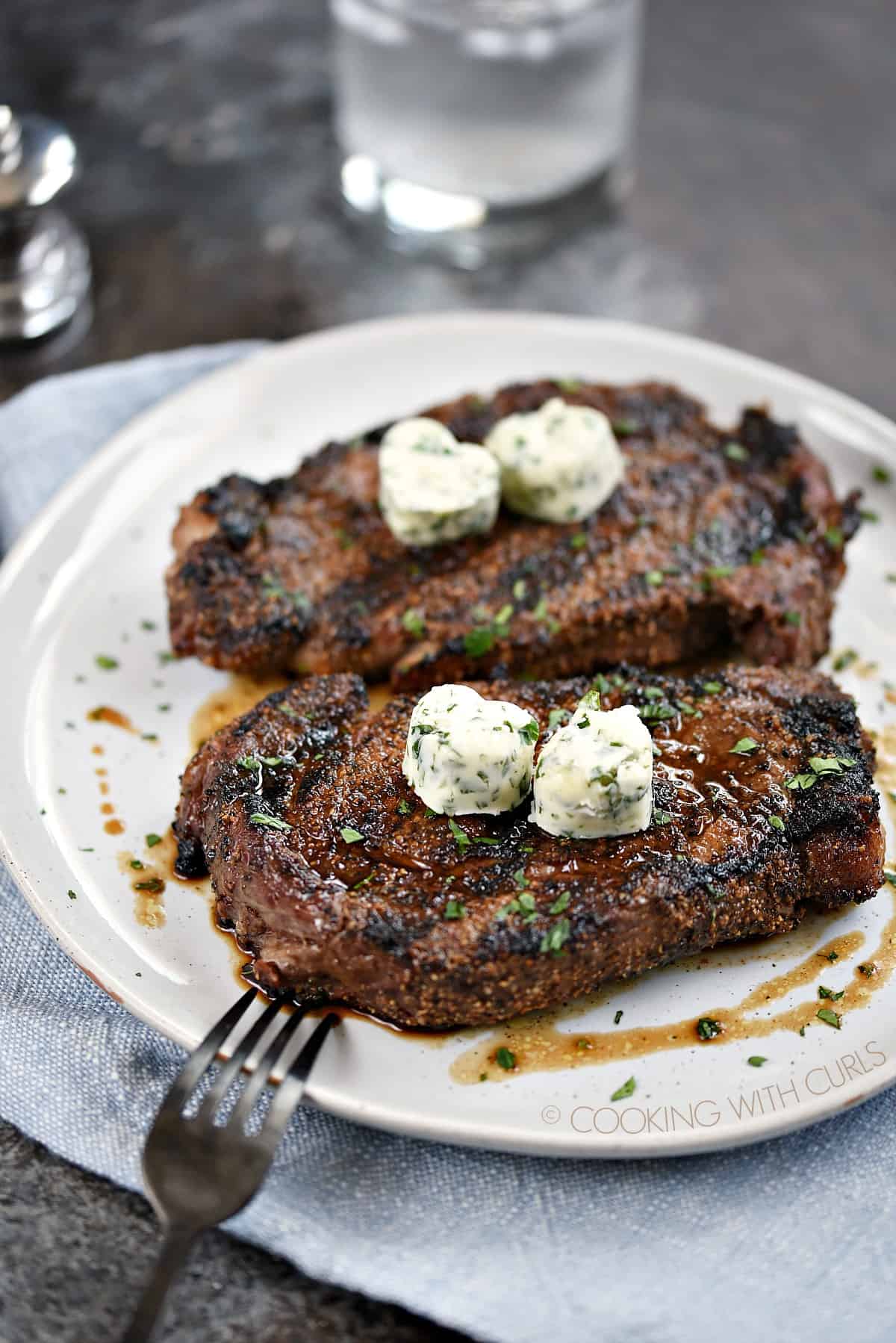 Two Perfect Grilled Steaks topped with two herb butter hearts sitting on a large white dinner plate.