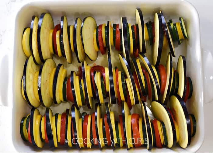 Zucchini, squash, tomatoes and eggplant slices lined up in a white baking dish. 